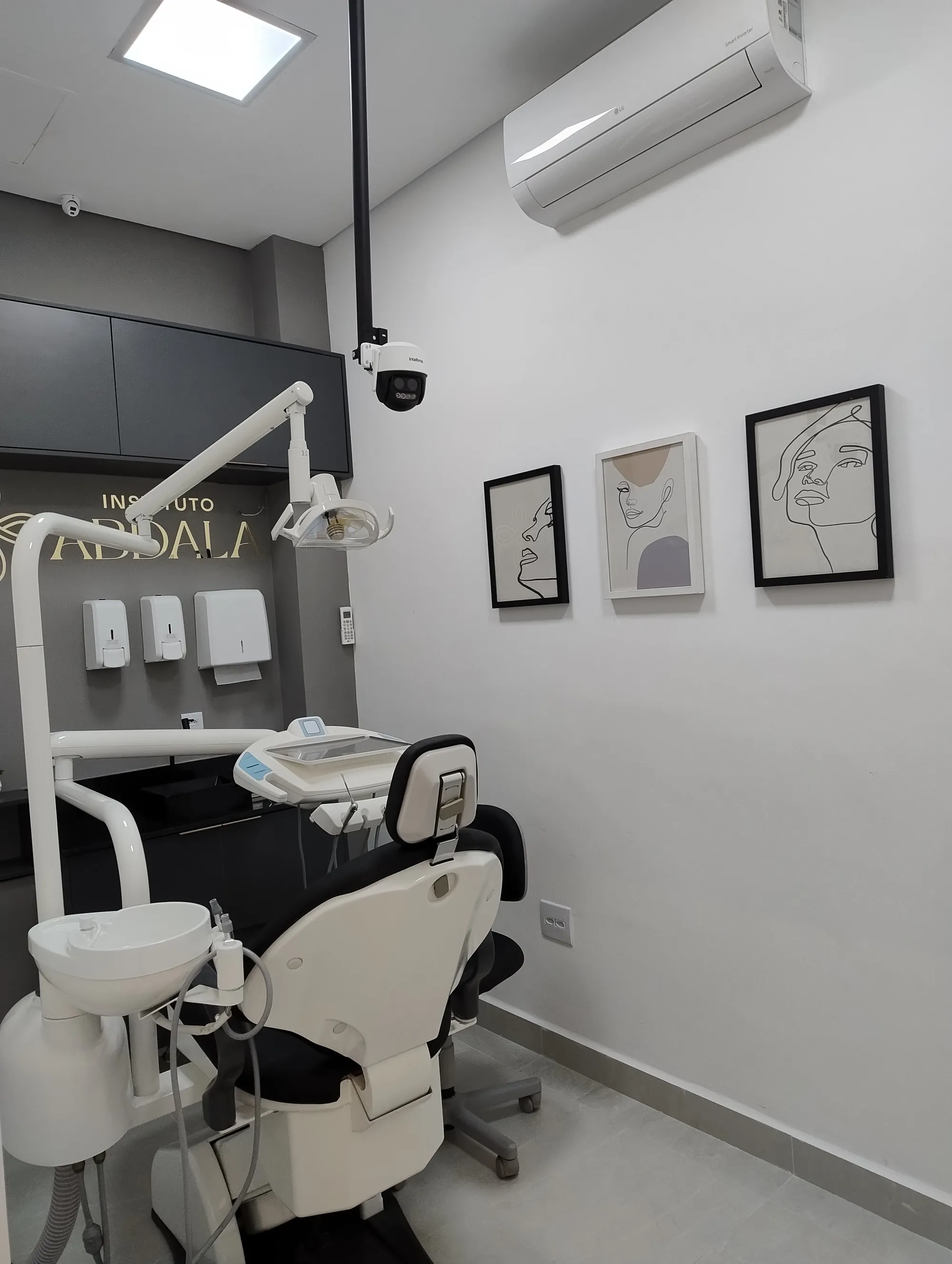 Consultório odontológico moderno com cadeira branca, equipamentos integrados, armários escuros e quadros com arte em linha.