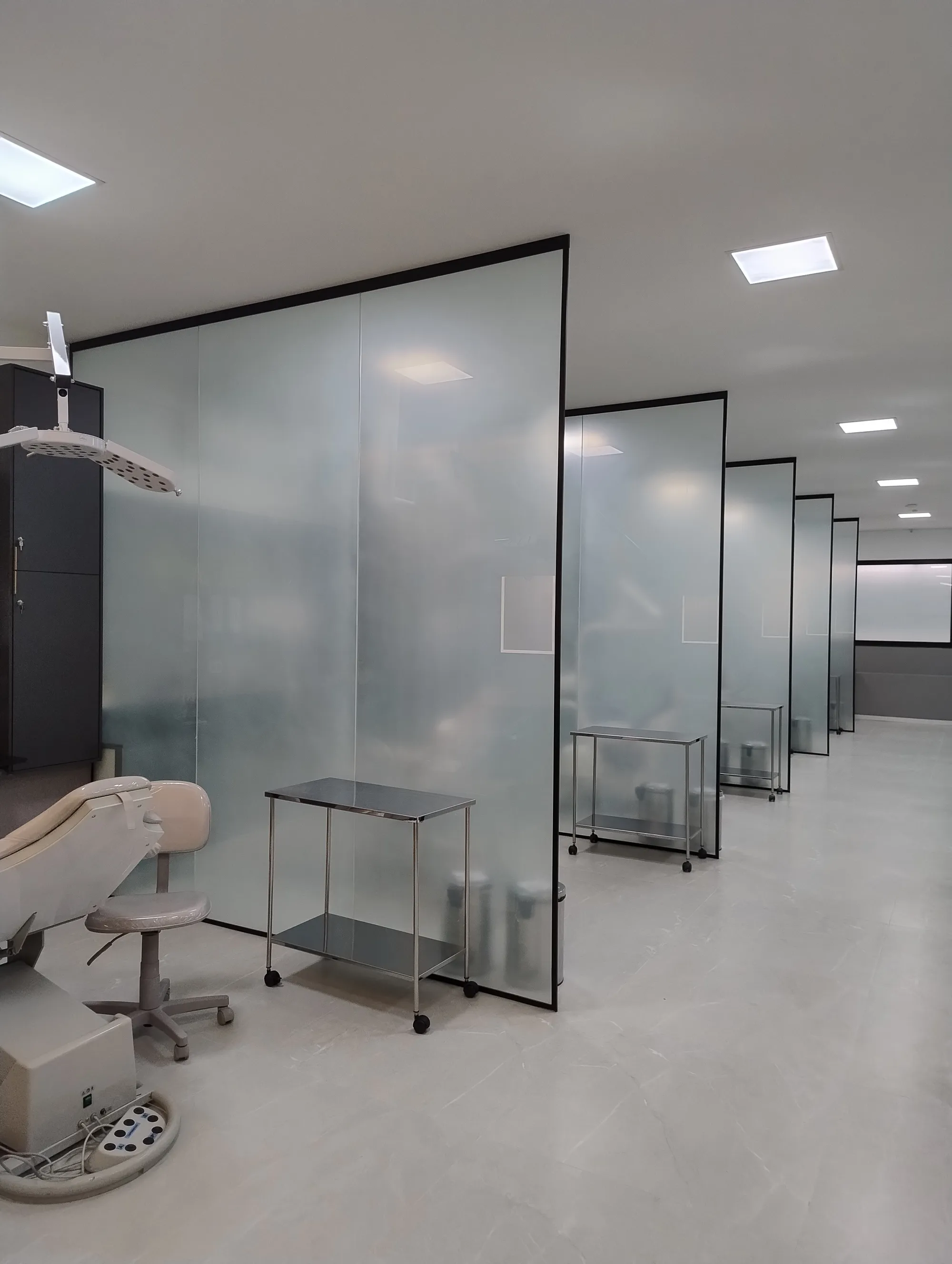 Salas de atendimento em conceito aberto, separadas por divisórias de vidro translúcido com estrutura preta, criando estações de trabalho contíguas.