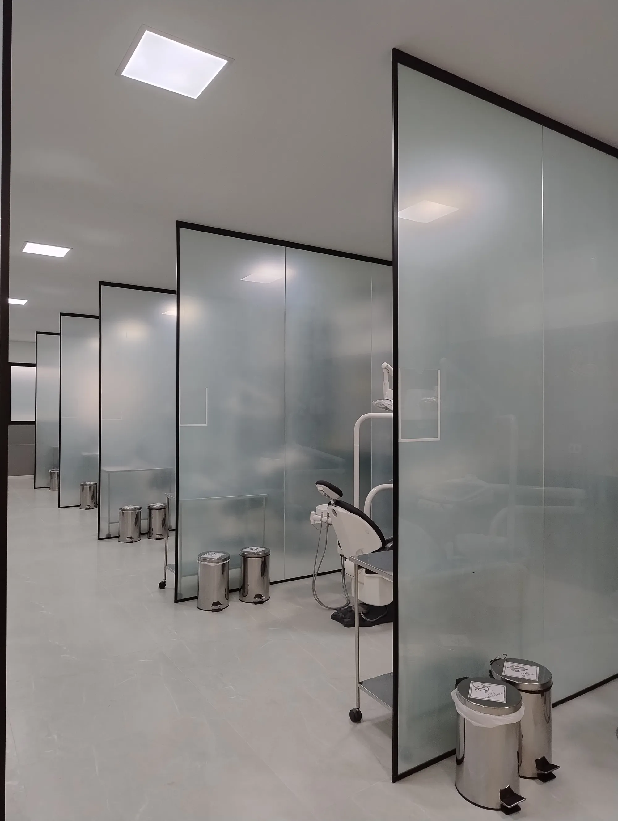 Corredor das estações de atendimento, mostrando a repetição das divisórias de vidro jateado e as cadeiras odontológicas perfiladas.
