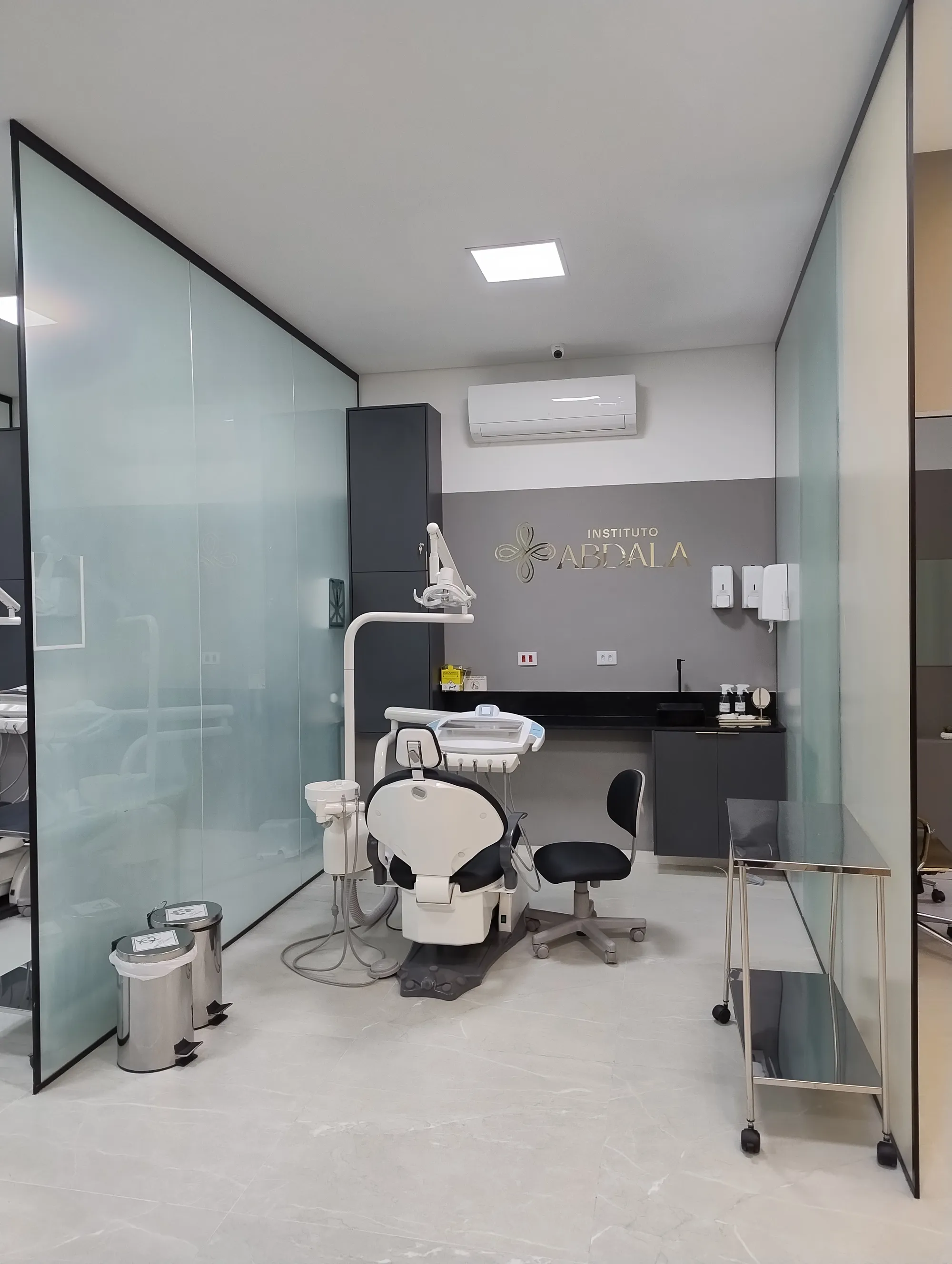 Interior de uma estação de atendimento odontológico, com cadeira principal, bancada de apoio, divisórias de vidro e logotipo iluminado na parede do fundo.