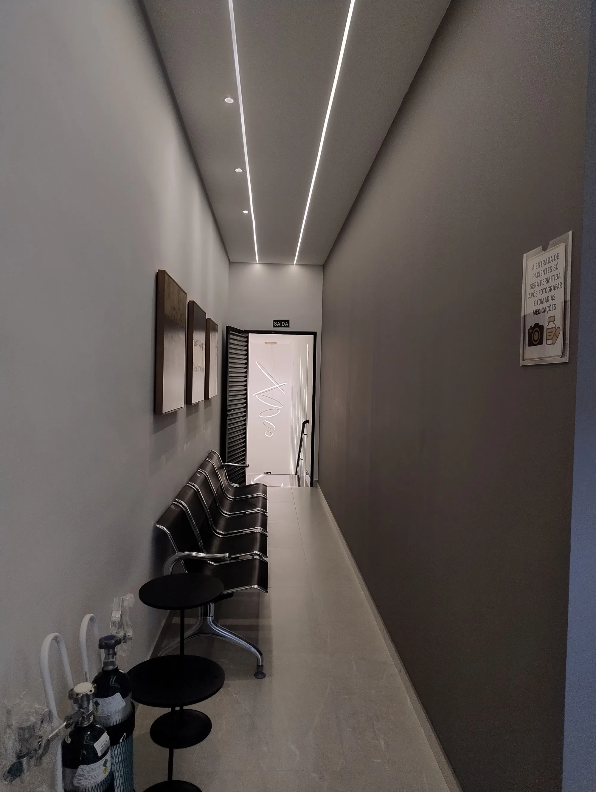 Corredor estreito de circulação, paredes em tom cinza escuro, com cadeiras de espera metálicas pretas dispostas linearmente sob iluminação contínua no teto.
