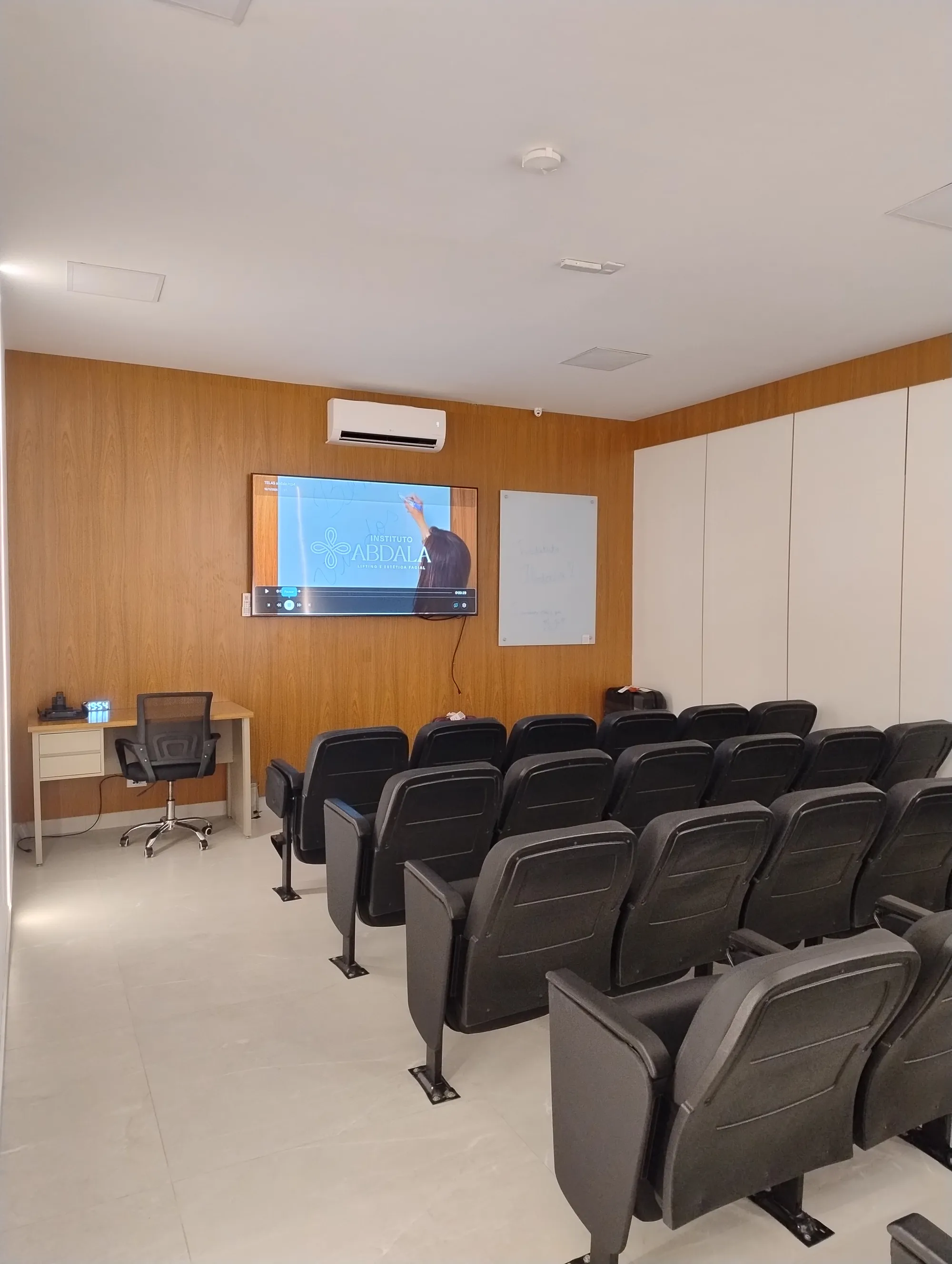 Sala de aula com painel frontal em madeira, TV com o logo do instituto, quadro branco e fileiras de cadeiras pretas com prancheta.