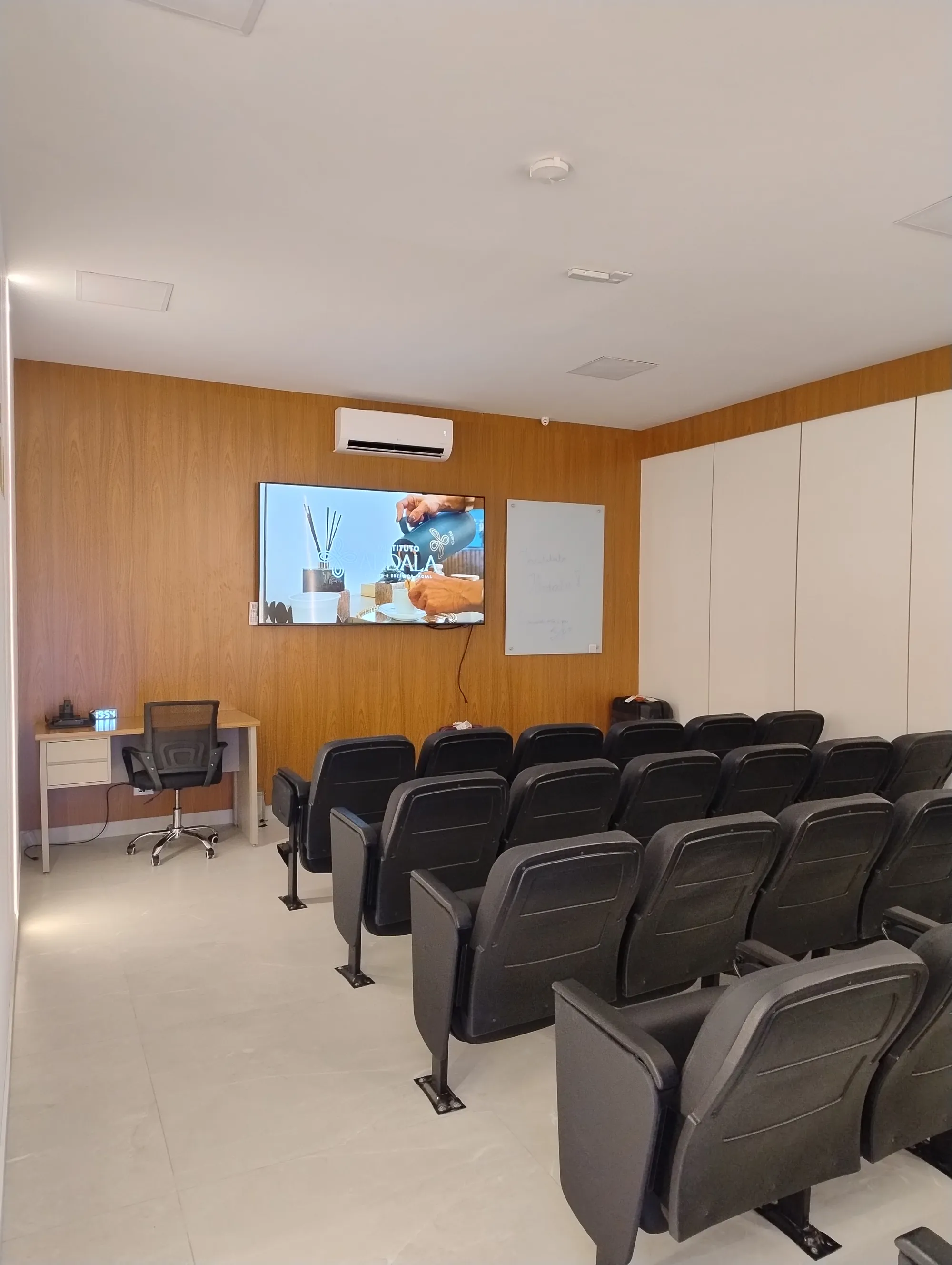 Outro ângulo da sala de aula, mostrando a TV ligada, mesa de apoio lateral para o professor e as fileiras de cadeiras para os alunos.