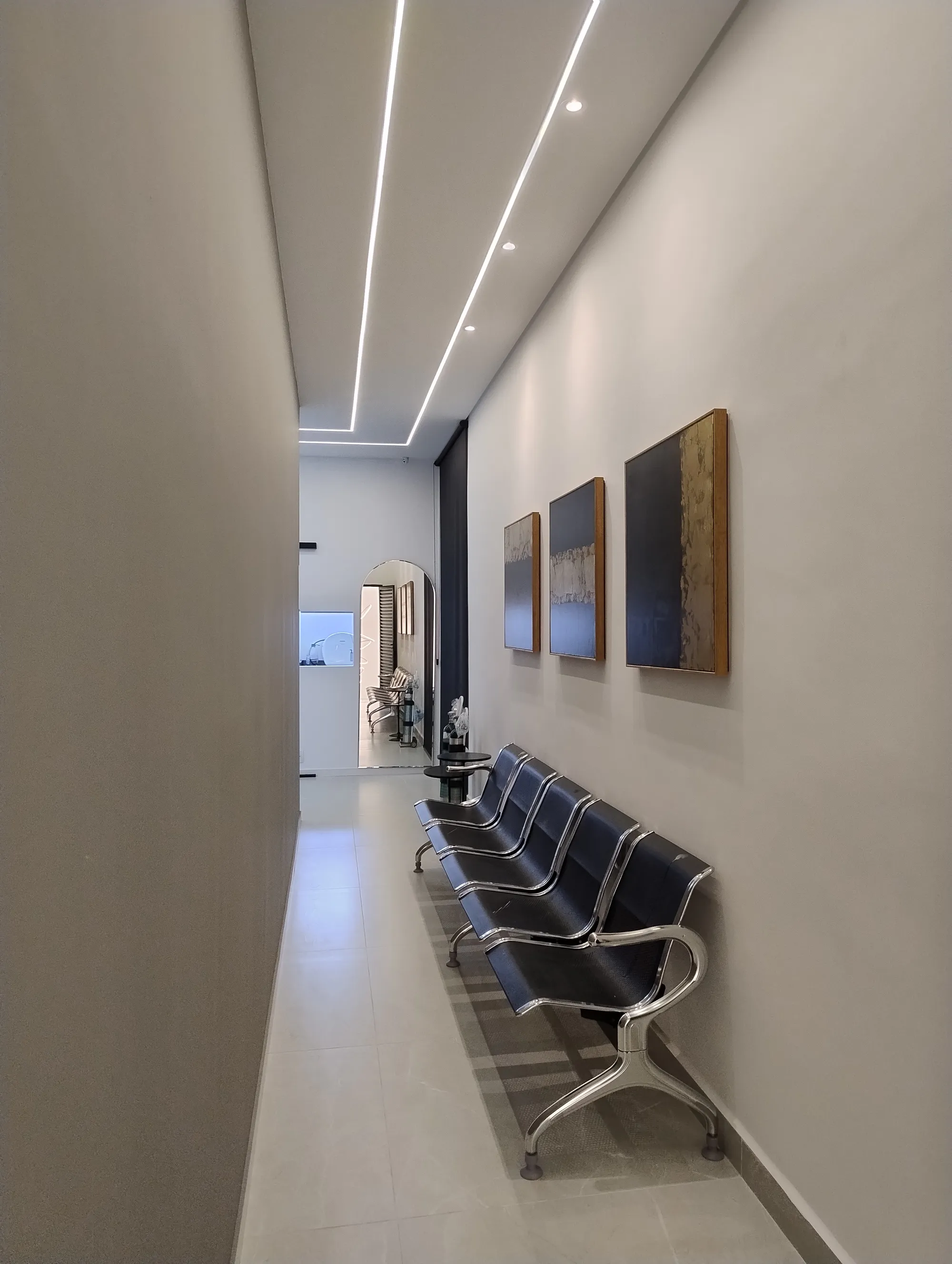 Corredor com paredes em cinza claro, três quadros alinhados na parede direita, iluminação embutida no teto em gesso rebaixado e cadeiras de espera metálicas.